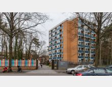 Mehrstöckiges Wohngebäude mit orangen Backsteinwänden und blauen Balkonen, umgeben von Bäumen. Im Vordergrund befindet sich ein bunter Zaun, zwei geparkte Motorroller, mehrere Autos und ein Fußweg, der in eine umzäunte Grünfläche führt.