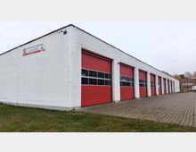 Flaches Feuerwehrgebäude mit weißer Fassade und mehreren roten Toren, teilweise mit Fensterstreifen. Davor gepflasterter Hof, seitlich Wiese, im Hintergrund ein weiteres Gebäude.