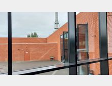 Blick durch große Fenster auf einen Hof mit roten Backsteinmauern, einer verschlossenen Metalltür und teilweise sichtbarer Begrünung. Im Hintergrund ein Industrieschornstein und bewölkter Himmel.