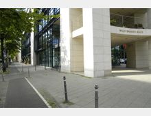 Moderne Gebäudefassade des Willy-Brandt-Hauses mit hellen Steinplatten und großflächigen Glasfronten. Im Vordergrund ein Geh- und Radweg, gesäumt von Pollern und Bäumen entlang der Straße.