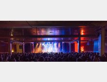 Ein Konzertsaal mit einer Bühne im Vordergrund, beleuchtet von bunten Scheinwerfern. Im Zuschauerbereich stehen viele Menschen vor der Bühne, und an der rechten Wand leuchtet ein Neon-Schild mit der Aufschrift „BAR“.