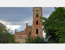 Eine alte Burgruine mit rotem Backstein und teils verfallener Fassade, bestehend aus einem hohen Turm mit mehreren großen, leeren Fenstern und einem niedrigeren Gebäudeteil mit Zinnen. Im Vordergrund sind Bäume und dichtes Grün zu sehen, der Himmel ist bewölkt.