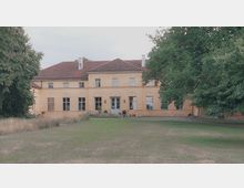 Gelbes, zweistöckiges Herrenhaus mit Satteldach, symmetrischen Fenstern und zentralem Eingang, umgeben von einer gepflegten Rasenfläche und mehreren großen Bäumen.