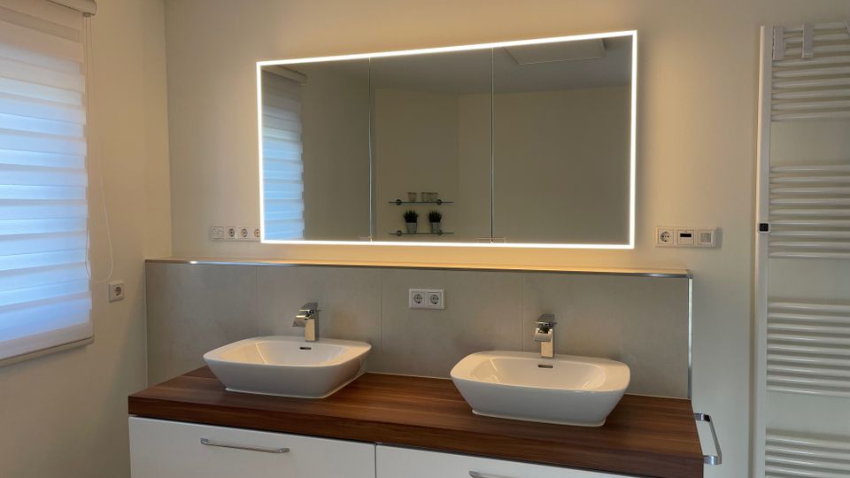 Modernes Badezimmer mit zwei rechteckigen Waschbecken auf einer hölzernen Arbeitsplatte, darunter weiße Schubladen. Ein großer, beleuchteter Spiegel hängt an der Wand, daneben ein weißer Handtuchheizkörper. Rechts und links sind weiße Wände mit schlichtem Dekor.