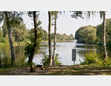 Flusslandschaft mit klarem Wasser, umgeben von dichtem Baumbewuchs. Im Vordergrund stehen Birken an einem Wiesenrand, daneben ein Schild mit Symbolen für Camping und Parkmöglichkeiten; im Hintergrund ist eine kleine Brücke erkennbar.