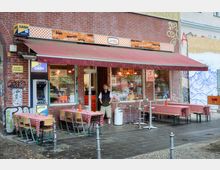 Außenansicht eines kleinen Imbisses mit Backsteinfassade, einer rot-weißen Markise und einem Geldautomaten an der Wand. Vor dem Geschäft stehen Tische und Stühle mit rot-weiß karierten Tischdecken auf dem Bürgersteig, im Hintergrund sind Graffiti an den Wänden zu erkennen.