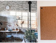 Loftartiger Raum mit rustikaler Backsteinwand, einem einfachen Schreibtisch mit Schreibmaschine, einem Holzstuhl und Dekoration wie Trockenblumen in einer Vase. Im Vordergrund eine große Korkpinnwand und eine Pflanze, links eine schlichte Tischlampe.