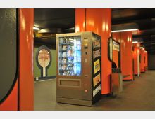 Ein U-Bahnhof mit orangefarbenen Säulen und abgerundeter Decke, ein Snackautomat steht prominent im Vordergrund. Im Hintergrund sind ein Wandbild und ein Stationsschild mit der Aufschrift „Siebenstern“ zu erkennen.
