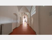 Langer Flur mit weiß gestrichenen Wänden und rotem Boden, gewölbter Decke und mehreren hängenden Laternen. Auf der rechten Seite befinden sich Türen und große Fenster mit Glasbausteinen, links steht ein Heizkörper.