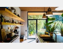 Moderne Küche mit großen bodentiefen Glastüren, die den Blick auf einen grünen Garten freigeben. Holzregale, Küchenutensilien und Pflanzen bestimmen den Raum, der durch natürliche und künstliche Beleuchtung hell und einladend wirkt.