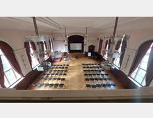 Ein historischer Saal mit hohen Decken, großen Fenstern mit roten Vorhängen und einem Parkettboden. Der Raum ist mit Reihen von schwarzen Stühlen bestuhlt, im Hintergrund gibt es eine Bühne mit einer Projektionsleinwand sowie einige Werbebanner und dekorative Elemente.