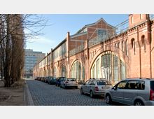 Straßenansicht eines alten, renovierten Backsteingebäudes mit großen, bogenförmigen Fenstern und einem Gittervorbau. Rechts parken Autos entlang einer gepflasterten Straße, links stehen schlanke, kahle Bäume.