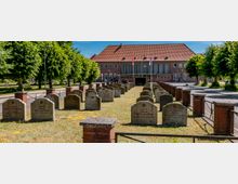 Reihen von Grabsteinen auf einer rechteckigen, von niedrigen Ziegelpfeilern und Metallgeländern eingefassten Grünfläche, flankiert von Bäumen. Im Hintergrund steht ein großes Backsteingebäude mit rotem Ziegeldach, großen Fenstern und Flaggenmasten.
