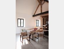 Ein heller, modern eingerichteter Raum mit Sprossenfenstern, sichtbaren Holzbalken an der Decke und poliertem Betonboden. Im Vordergrund stehen gepolsterte Holzstühle, im Hintergrund ein langer Holztisch mit verschiedenen Stühlen und ein offenes Regal an der Wand.