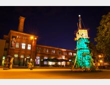 Backsteingebäude mit einem Schornstein im Hintergrund, beleuchtet bei Nacht, neben einer bunt illuminierten, kunstvollen Installation mit Pflanzen. Im Vordergrund stehen Bänke und Sonnenschirme auf einem offenen Platz.