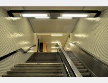 Treppenaufgang und Rolltreppe in einer U-Bahn-Station mit weißen gefliesten Wänden, metallischen Geländern und Deckenbeleuchtung. Die Umgebung wirkt funktional und schlicht gestaltet.