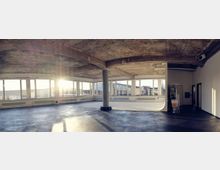 Ein leerer, loftartiger Raum mit roh belassener Betondecke, großen Panoramafenstern und einem grauen Boden. In der linken Raumecke fällt Sonnenlicht durch die Fenster, während in der rechten Ecke einige Stative und Geräte stehen.