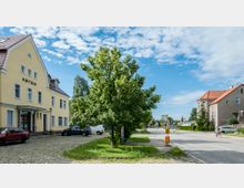 Straßenszene in einer Kleinstadt mit einem gelben Gebäude mit der Aufschrift „Ketzin“ links, einem Baum in der Mitte und Wohngebäuden mit roten Dächern rechts. Pflastersteine, Grünstreifen und parkende Autos prägen den Vordergrund, während eine Bushaltestelle und ein Radfahrer am rechten Bildrand sichtbar sind.