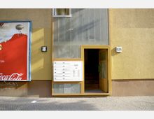 Gelbe Hausfassade mit einer gläsernen Eingangstür, daneben eine Briefkastenanlage und die Hausnummer "63A". Links befindet sich ein Werbeplakat mit rotem Hintergrund. Der Gehweg davor ist mit Pflastersteinen belegt.