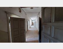 Ein alter, verlassener Flur mit offenen Holztüren auf beiden Seiten. Die Wände sind weiß gestrichen, weisen aber Schäden und freiliegende Kabel auf; der Boden besteht aus abgenutztem Holz.