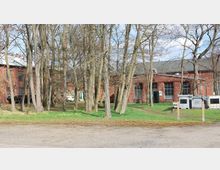 Backsteinbau mit großen Fenstern und Satteldach, umgeben von kahlen Bäumen auf einer Grünfläche. Im Vordergrund befindet sich ein unbefestigter Weg sowie gestapelte Container.