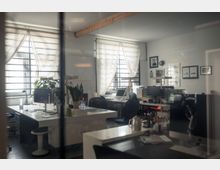 Ein modernes, kleines Büro mit mehreren Schreibtischen, Monitoren und Bürostühlen. Die Fenster haben halb geöffnete weiße Vorhänge, es sind Pflanzen, Wandbilder und Dekorationen sichtbar, während Tageslicht in den Raum fällt.