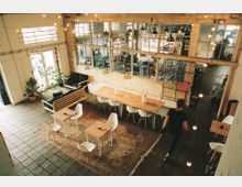 Industrie-inspiriertes Café mit großzügigem Raum, Holztischen, weißen Stühlen und einem großen Teppich. Eine Glaswand trennt den Sitzbereich vom hinteren Werkstattbereich, während Lichterketten von der Decke hängen.