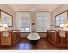 Elegantes Badezimmer mit freistehender Badewanne in der Mitte, flankiert von zwei Waschtischen mit Holzverkleidung und großen Spiegeln. Im Hintergrund drei große Fenster mit halb-transparenten Rollos und einem kleinen Bild an der Wand, darunter ein Heizkörper und ein Holzboden.
