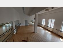 Großer Tanz- oder Übungsraum mit hellem Parkettboden, Spiegelwand, Ballettstangen und mehreren Fenstern, die Tageslicht hereinlassen. An den Wänden befinden sich Heizkörper, und ein Metalltür ist an der Stirnwand sichtbar.