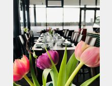 Ein modern eingerichteter Restaurantbereich mit einem langen, gedeckten Tisch, dekoriert mit Geschirr und Blumenarrangements. Im Vordergrund sind bunte Tulpen zu sehen, während große Fenster im Hintergrund Tageslicht hereinlassen.