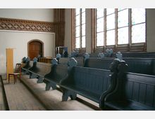 Innenraum einer historischen Kirche mit dunklen, hölzernen Kirchenbänken, farbigen Bleiglasfenstern und einem gemauerten roten Zierband an der Wand. Im Vordergrund stehen ein Holzpult und ein einfacher Stuhl auf einem Holzfußboden.