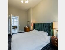 Ein Schlafzimmer mit einem großen Bett, dessen Kopfteil gepolstert und grün ist. Im Hintergrund führt eine offene Tür zu einem weiteren Raum mit einem großen Fenster, durch das Tageslicht einfällt.