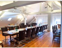 Großer Konferenzraum mit Holzboden und sichtbaren weißen Dachbalken unter einer Dachschräge. Der Raum ist mit langen Holztischen und gepolsterten Stühlen eingerichtet, auf den Tischen stehen Wasserflaschen; im Hintergrund befinden sich ein Buffet mit Getränken und ein Sitzbereich mit Sesseln.