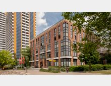 Moderne Backsteinfassade eines mehrstöckigen Büro- oder Verwaltungsgebäudes mit großen Glasfenstern. Im Hintergrund befinden sich hohe Wohngebäude, während Bäume und Flaggen im Vordergrund den Bereich säumen.