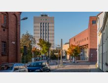 Städtische Straße mit Kopfsteinpflaster, flankiert von Backstein- und Putzgebäuden. Im Hintergrund ein grauer Hochhausblock und ein modernes Backsteingebäude; Bäume mit herbstlichem Laub säumen die Straße.