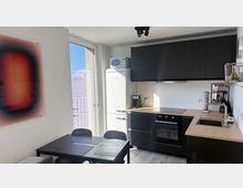 Moderne, kompakte Wohnküche mit schwarzen Hängeschränken, Holzarbeitsplatte, Herd mit Backofen und Spülbecken sowie freistehendem Retro-Kühlschrank. Daneben ein Esstisch mit Stühlen vor einer großen Balkontür, die viel Tageslicht hineinlässt.