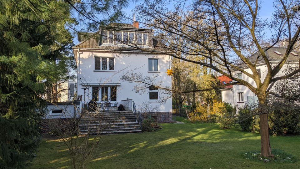 Rückansicht eines weißen, mehrstöckigen Wohnhauses mit schrägem Ziegeldach und Dachgauben, davor eine breite Treppe zur Terrasse. Im Vordergrund liegt ein großer Rasen mit Bäumen, Sträuchern und einem kleinen Holzstapel, nebenan ist ein weiteres Wohnhaus zu sehen.