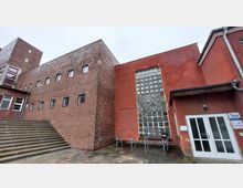 Backstein-Gebäudefassade mit kleinen rechteckigen Fenstern, neben einem roten Gebäudeteil mit einer großen Glasbausteinwand. Im Vordergrund führen steinerne Treppenstufen auf einen gepflasterten Platz, auf dem Fahrradständer und eine Tür mit Glasfenstern sichtbar sind.