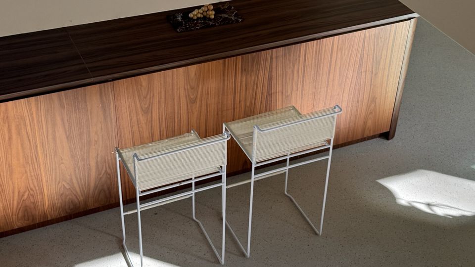 Minimalist interior corner with a long wood-veneer sideboard against a plain wall and two small metal-framed stools in front. Sunlight casts large rectangular patches of light and shadow across a smooth gray floor.