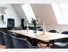 Modern eingerichteter Besprechungsraum mit einem Holztisch, umgeben von gepolsterten, grauen Stühlen. Auf dem Tisch stehen eine Vase mit Blumen und zwei weiße Kerzenhalter; im Hintergrund fällt Tageslicht durch große Dachfenster ein.