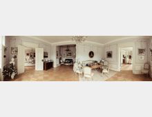 Ein eleganter historischer Salon mit verzierten weißen Wandpaneelen, Stuckarbeiten und Kronleuchtern. Der Raum ist mit antiken Möbeln, einem Holzboden und dekorativen Kunstwerken geschmackvoll eingerichtet; geöffnete doppelflügelige Türen führen in angrenzende Räume.