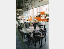 Modernes Café mit einer offenen, hellen Inneneinrichtung. Dunkle Holztische und -stühle sind mit Gläsern, Besteck und Blumenvasen gedeckt, während orangefarbene Regale und Pflanzenakzente den Raum dekorieren; große Fenster lassen Tageslicht hinein.