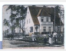 Historisches Bild eines zweigeschossigen Gebäudes mit Satteldach, umgeben von Kiefern und eingezäuntem Garten. Vor dem Haus stehen mehrere Personen, darunter Erwachsene und Kinder, auf einem Gehweg an einer unbefestigten Straße.