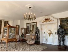 Wohnzimmer im historischen Stil mit antikem Mobiliar und dekorativen Elementen. Zu sehen sind eine Vitrine mit Porzellan, eine Polster-Sitzgruppe, Ritterrüstungen, ein Kronleuchter, eine weiße Tür mit goldenen Verzierungen und Wandteppiche.