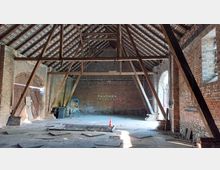 Innenraum eines alten Scheunengebäudes mit freiliegendem Dachgebälk und Wänden aus rotem Ziegelstein. Auf dem Boden liegen Schutt und ein Gitterrost, an der Wand steht „Rauchen verboten“, und durch eine offene Holztür fällt Licht ein.