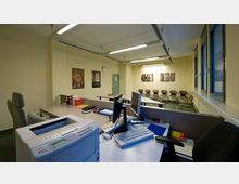 Großzügiger Büroraum mit Empfangsbereich, ausgestattet mit Schreibtischen, Computern und einem Drucker im Vordergrund. Im Hintergrund befinden sich eine Sitzgruppe mit Stühlen, eine Garderobe und gerahmte Bilder an den hellgelben Wänden; auf der rechten Seite verläuft eine Fensterreihe.