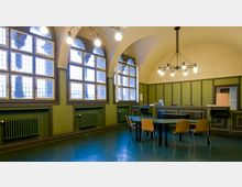 Innenraum eines historischen Saals mit einem gewölbten Deckenbereich, großen Sprossenfenstern und grün gestrichener Wandvertäfelung. In der Mitte stehen ein ovaler Tisch und Stühle, während im Hintergrund ein Empfangstresen sichtbar ist.