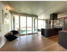 Moderner Wohnraum mit offener Küche, großen Fenstern und Blick auf einen Balkon mit Pflanzen. Die Küche ist in dunklen Farben gehalten, mit einer zentralen Insel, während der Boden aus hellem Holz besteht.