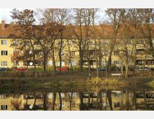 Wohngebäude im Hintergrund mit gelber Fassade und roten Ziegeldächern, dahinter eine Baumreihe ohne Laub. Im Vordergrund ein kleiner Teich, der die Szenerie reflektiert, sowie eine Wiese mit einer Sitzbank.