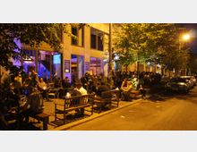 Eine belebte Straßenszene bei Nacht mit Menschen, die vor einem Gebäude mit großen Fenstern auf Bänken und Stühlen sitzen. Innen sind bunt beleuchtete Räume zu sehen, während Bäume und geparkte Autos die Straße säumen.